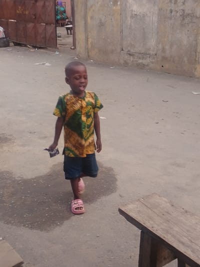 A young child is walking in a street. The child is wearing a patterned shirt with green and brown colors, denim shorts, and pink sandals. In their left hand, they hold an object. The street appears to be in an urban area, with buildings and gates visible in the background. A wooden bench is partially visible on the right side of the image. The street surface is concrete and appears to be wet in places.
