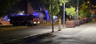 The image depicts a dark urban street scene in Huesca, Spain, at night. A red fire truck with blue flashing lights is parked on the street, facing the viewer's right. The fire truck has "BOMBEROS" written on its front in reverse. Adjacent to the street is a sidewalk paved with light-colored square tiles, illuminated by streetlights and the fire truck's lights. The sidewalk is lined with large, rectangular planters made of rusted metal, each containing greenery. Several trees with lush green foliage are present, casting shadows on the street and sidewalk. In the background, a multi-story brick building is visible, with illuminated windows. There is a traffic barrier and a street light pole on the sidewalk. The overall impression is one of an emergency response or a temporary situation on a quiet city street at night.