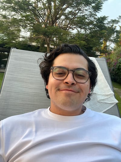 A man with dark hair and glasses is reclining on a light gray chaise lounge. He is wearing a white t-shirt. A lush green tree is visible in the background to the left. The photo appears to be taken outdoors in a garden or yard setting. The lighting is soft and natural, suggesting it is daytime.