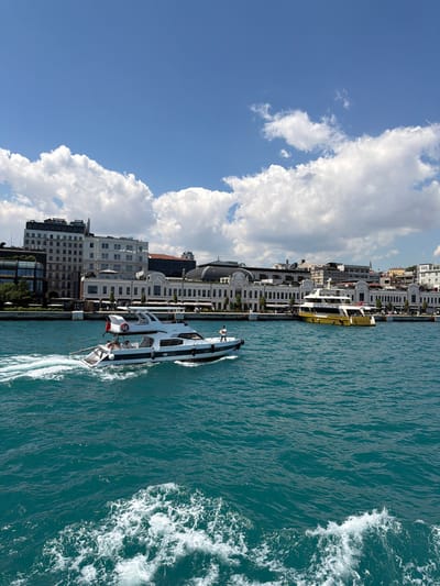 This image captures a bright, sunny day with a clear blue sky dotted with fluffy white clouds. The scene overlooks a vibrant turquoise body of water, likely the Bosphorus Strait, with a shoreline in the background featuring several modern buildings.

In the foreground, a white yacht with a blue stripe is moving at a moderate speed, creating white wakes in the water. Several people are visible on the yacht. To the right of the yacht, a larger, yellow and white ferry is docked along the shore.

The shoreline itself is lined with a long, elegant white building with arched windows and a grand facade. Behind this structure, modern office buildings rise, including one with the "JW Marriott" sign prominently displayed. The overall impression is a bustling urban waterfront, suggesting a location in a prominent city. Given the architectural style and the presence of luxury hotels, this scene is consistent with the Karaköy neighborhood of Beyoğlu, Istanbul, Turkey, known for its historic buildings and modern developments along the Bosphorus.