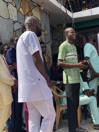 The image captures a group of Black males gathered outdoors in Kumasi, Ghana, under clear, sunny conditions. In the left foreground, a man with a bald head and beard, dressed in a white short-sleeved tunic and white trousers, stands in profile, looking towards the right side of the frame. To his immediate right, a man with a short haircut, wearing a green patterned short-sleeved shirt with distinct white and black horizontal stripes across the chest, and black trousers, is smiling widely towards the right. He holds a black smartphone in his left hand and a black bag in his right, while wearing dark sandals.
Behind these two individuals, other men are partially visible: one in a light cream-colored long-sleeved garment, another wearing a dark blue polo shirt and sunglasses, and a younger man in a dark shirt. To the right of the smiling man, another individual wearing a light green short-sleeved tunic and matching trousers is seated on a beige plastic chair.
The background consists of a building facade covered in irregular, multi-colored stone tiles, primarily grey, brown, and tan. Above the facade, an ornate black metal balcony railing is visible. Numerous tangled electrical wires traverse the upper portion of the frame against a bright blue sky. A small white security camera is mounted on the building wall to the left. The ground appears to be a paved or tiled surface.