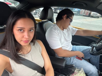 A man and a woman are in the front seats of a car. The woman is in the right seat, wearing a light-colored, button-down shirt with a seatbelt across her chest, and has dark hair. The man is in the left seat, wearing a white t-shirt and blue-framed glasses, and is reaching into his jeans pocket. There is a flower bouquet on his lap. A "Scotiabank" credit card is visible in his hand. The car is parked on a street in San Juan Tejaluca, Mexico, as indicated by the Spanish text on the "Scotiabank" card. The scene outside the car window shows buildings and vehicles.