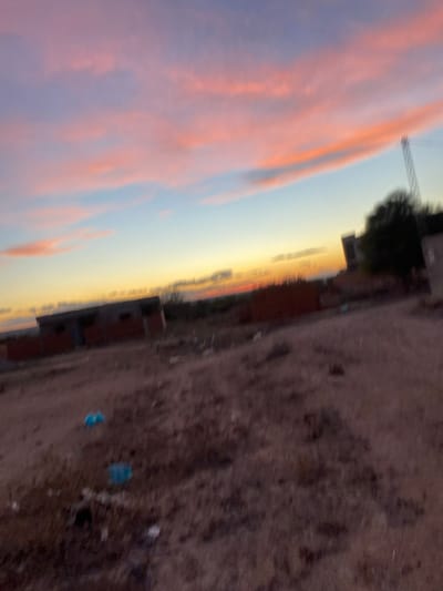 The image depicts an outdoor scene in الغنادة, Tunisia, at either sunrise or sunset, characterized by a vibrant sky. The upper portion of the frame features a sky with horizontal bands of color, including shades of pink and orange in the upper clouds, transitioning to lighter blue, and then to a yellowish-orange near the horizon. The clouds exhibit some blur, suggesting motion or photographic artifact.
The midground and foreground consist of an expanse of uneven, reddish-brown earth, appearing dry and sparsely vegetated. Scattered debris, including several distinct bright blue plastic objects and lighter-colored litter, is visible on the ground in the foreground.
In the left midground, there is an unfinished or derelict structure composed of dark-colored blocks or bricks, with multiple open rectangular spaces likely intended for windows or doors. To the right of the frame, another building or structure, also appearing dark, is partially visible, obscured by a dense, dark green tree. A tall, slender structure, possibly a communication tower, is faintly discernible behind the tree in the upper right quadrant. The overall image exhibits motion blur and low resolution. No individuals are present.