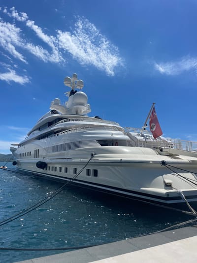 A large white luxury yacht named "Bellamus" is docked in the city of Tivat, Montenegro, on a sunny day with a blue sky and scattered white clouds. The yacht is prominently featured, taking up most of the frame. Its sleek, multi-deck structure is visible, with numerous windows lining its hull. The yacht is secured to a dock by thick black ropes. Two dark fenders are attached to the side of the yacht to prevent damage. A red flag with a crest and a Union Jack in the canton is flying from a mast on the yacht. The water around the yacht is a deep blue and shows some ripple effects from the sun. In the distance, a hazy green coastline is visible. The overall scene depicts a serene and opulent maritime setting.