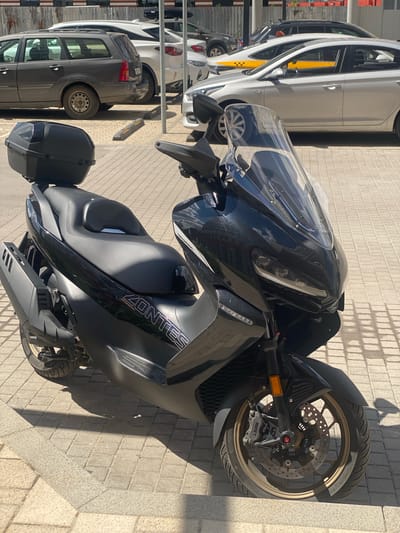 The image shows a black scooter parked on a sidewalk made of light gray paving stones. The scooter is a Zontes model, with "ZONTES" written in white lettering on its side. The scooter has a large clear windshield and black handlebars. The front wheel has an ABS system. To the left of the scooter, there are several parked cars, including a white taxi with a yellow stripe and black checkered pattern. The cars are parked in what appears to be a parking lot. The sun is casting shadows on the ground, suggesting it is daytime. The location is Kotelniki, Russia. There are no people visible in the image. The scene is still and quiet, with no specific activity taking place.