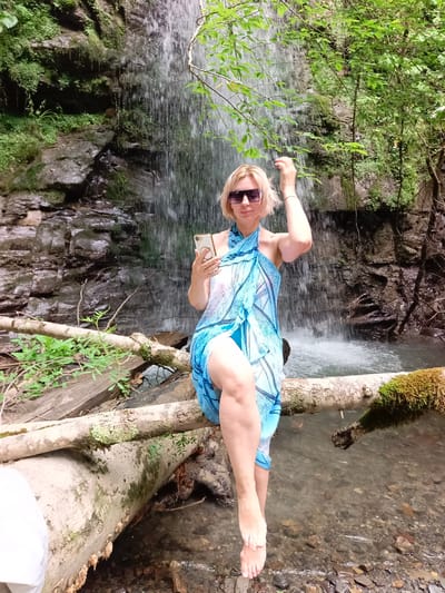 A blonde woman with sunglasses sits on a fallen tree trunk by a waterfall, holding a smartphone. She is wearing a blue sarong and has her right hand raised to her hair. The waterfall cascades down a rocky, moss-covered cliff face into a shallow stream. The surrounding area is dense with green foliage and trees, suggesting a natural, outdoor environment. The image was taken in Plastunka, Russia.