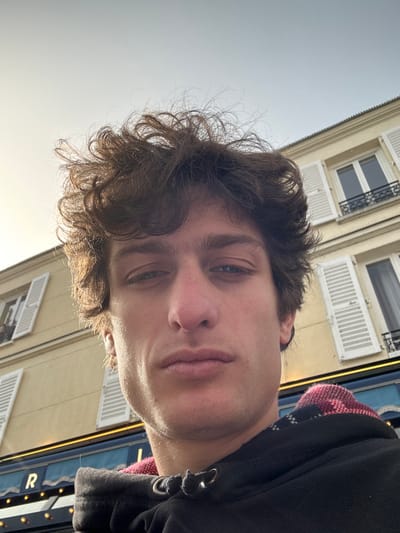 A single male subject is depicted in a close-up, upward-angled self-portrait in Vincennes, France. The subject, appearing as a young adult male, possesses curly, disheveled brown hair, dark eyebrows, and light-colored eyes, and maintains a neutral direct gaze towards the camera. He is dressed in a black hooded sweatshirt, with a visible red and black plaid pattern lining or garment underneath the collar area. The background features a multi-story building with a light-colored facade, likely beige or yellow, under a bright, partially overcast sky. Architectural details include white louvered shutters on the windows and black wrought iron balustrades. Beneath the residential floors, a commercial storefront is visible, distinguished by a blue awning with white illuminated lettering, where the letter "R" is distinctly discernible. The scene is illuminated by natural daylight, creating subtle shadows on the subject's face.