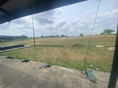 This image depicts a wide view of a sports field, likely the Uyo Township Stadium in Uyo, Akwa Ibom State, Nigeria, observed from a covered grandstand area.
In the immediate foreground, the concrete steps of a spectator seating area are visible, featuring dark green painted risers. These steps show signs of wear and contain scattered white plastic litter, including bottles and wrappers. Above, a corrugated metal roof extends over the viewing position. A tall, thin pole, painted blue and white, stands vertically near the edge of the concrete steps, extending upwards out of frame.
The midground is dominated by a large, open field, primarily covered in dry, brown grass interspersed with patches of green. A small mound of dark earth or debris is situated on the field to the right of the blue pole. Approximately six human figures are visible in the middle distance on the field; their specific actions are indistinct due to distance, but they appear to be standing or moving, possibly engaged in recreational activity.
In the background, beyond the field, indistinct buildings and a sparse line of trees are visible on the horizon. A series of taller, dark poles, potentially floodlights or goalposts, are discernible along the left perimeter of the field. The sky is heavily overcast, filled with various shades of grey and white clouds, indicating a cloudy or partly cloudy daytime setting. The overall scene suggests an active public sports or recreational facility with evident signs of use and some environmental disarray from litter.