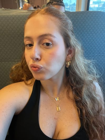 A close-up image depicts a female individual with long, wavy, reddish-brown hair, light skin, and visible freckles on her chest and shoulders. She wears a black halter-neck top, multiple gold-colored earrings in her right ear (including a large hoop, a smaller hoop, and a stud), and two layered gold-colored necklaces—one with a white butterfly pendant and another with a rectangular 'E' initial pendant. Her lips are pursed, and her eyes gaze directly at the camera. A slight reddish mark is visible on her left cheek. The background features a dark gray, patterned upholstered seat, consistent with public transit. Through a window on the upper right, a blurred daytime view of outdoor structures and sky is visible. The top of another person's head, showing braided hair, is partially visible in the seat ahead. The scene takes place on a public transit vehicle in Linden, United States.