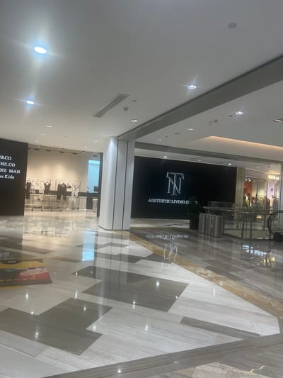 The image depicts an interior view of a modern shopping mall in Liandu District, China. The scene is well-lit with a combination of recessed ceiling lights and ambient mall lighting. The flooring is a polished marble or similar stone, laid in a geometric pattern of light and dark grey and white tiles, with a golden accent stripe running through it. The reflective surface of the floor mirrors the lights and structures above.
On the left side of the image, a store's dark facade displays text, partially visible, including what appears to be "&CO", "NZ.CO", "NZ MAN", and "oz Kids". In front of this store, racks of clothing are displayed on a white tiled floor. The clothing items are predominantly black and white, appearing to be fashion garments.
To the right, a large, black wall features a stylized logo and the text "AESTHETIC LIVING SPACE" in white, illuminated lettering. Adjacent to this wall are three silver cylindrical trash receptacles. Further to the right, a section of the mall shows store windows with clothing displays and escalators leading upwards, suggesting multiple levels. The overall environment is clean, spacious, and designed for retail shopping. There are no visible people in the immediate foreground, though there may be some indistinct figures within the storefronts on the right.