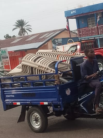 In Pantame, Nigeria, a blue three-wheeled vehicle, identified by a license plate that reads "GMJ 1738," transports a stack of beige plastic chairs. The vehicle is being driven by a young man wearing a navy blue shirt and dark pants, his right hand on the steering mechanism. Behind the vehicle, a red truck is visible, and further in the background, buildings with corrugated metal roofs are present. One building is painted blue with the letters "APC" prominently displayed on its facade, and another building features a red sign with the word "Itafoam." A palm tree stands tall against a cloudy sky.