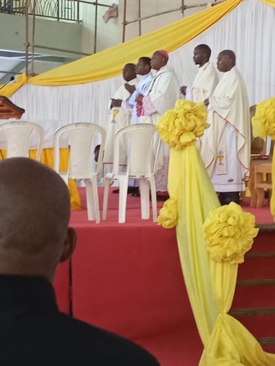 The image shows a scene inside a building in Arusha, Tanzania, at coordinates -3.372, 36.692. There are four men, likely priests, standing on a stage. They are wearing white robes with gold trim. One priest is wearing a red hat. The stage is decorated with white and yellow fabric. The men are standing in front of a table with chairs behind them. The stage is elevated and has a red carpet covering it.  There is a man standing in the foreground, his face obscured, with a black shirt. The room is decorated with yellow fabric and flowers.