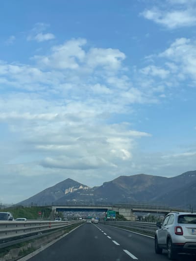 The image displays a multi-lane highway scene in Scafati, Italy, under a partly cloudy sky with mountainous terrain in the background. The perspective is from within a vehicle moving forward on the highway.

In the foreground, the rear of a white SUV, possibly a Fiat 500X, is visible on the right, showing a partial license plate beginning with "GC". The asphalt road features white dashed lane markings, delineating multiple lanes of traffic. To the left, a metal guardrail separates the highway lanes, with other white vehicles discernible in a parallel lane further back.

The midground shows moderate traffic on the highway. An elevated concrete overpass with blue metal support structures spans horizontally across the highway. Beyond this overpass, a green highway sign on the right indicates routes to "NOCERA" and "SALERNO". A distant urban landscape is visible beneath the bridge.

The background is dominated by a range of dark, rugged mountains, characterized by both vegetated slopes and exposed rock faces. The sky is largely blue, interspersed with numerous white and grey cumulus clouds. No specific individuals or interactions are identifiable.