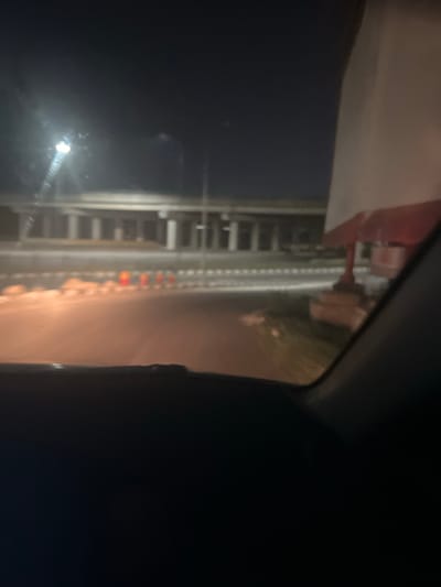 The image captures a nighttime scene in Aso, Nigeria, from the interior of a vehicle, looking out at a brightly lit structure. The primary subject is a section of what appears to be a roller coaster track or a similar amusement park ride. The track is constructed from metal supports and runs horizontally across the frame.

Along the track, a series of small, distinct lights, predominantly red, orange, and yellow, illuminate parts of the structure, suggesting decorative lighting or operational indicators for the ride. A prominent, bright light source, possibly a headlight or a streetlamp, is visible towards the left side of the frame, casting a significant glare or reflection on the vehicle's window.

The foreground and background are mostly shrouded in darkness, indicating the late hour. Parts of the vehicle's interior, including a dark dashboard or window frame, are visible on the right, framing the view. There are no people visible, and no discernible text. The scene evokes the atmosphere of an amusement park or fairground at night.