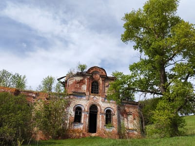 The image shows the ruined Church of the Life-Giving Trinity in the village of Крынды, Russia. The building is constructed of red brick and is heavily weathered, with missing sections of the roof and walls. Vegetation is growing on and around the structure. A large tree stands to the right of the building, partially obscuring the view. The sky is overcast with some patches of blue visible. The ground is covered in green grass.