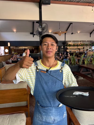 An indoor-outdoor dining establishment in Pecatu, Indonesia, is depicted. A male individual, appearing to be a server, occupies the foreground, facing the camera with a smile. He wears a yellow and white striped t-shirt under a denim bib apron secured with brass-colored buckles; a small brown patch is affixed to the apron's bib, and a wave-like logo is present on the lower right. A black wired earbud is inserted into his left ear, with the wire extending downwards. A silver-toned bracelet adorns his right wrist, and he holds a black serving tray in his left hand, which contains a crumpled white paper item. His right hand is raised, forming a 'thumbs up' gesture. A pen is visible in a pocket on his apron. The background reveals a spacious restaurant with high, angled ceilings featuring exposed dark beams, white paneling, multiple ceiling fans, and woven light fixtures. Seating arrangements include upholstered wooden chairs and booths with light cushions on the left, and wooden picnic-style tables and benches on the right, separated by planters containing snake plants. A bar area with bottles and hanging plants is visible in the mid-ground. Multiple patrons are seated throughout the establishment. The environment is brightly lit, suggesting daylight.