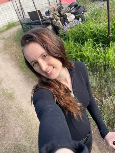 The image depicts a selfie of an adult female, positioned outdoors in a semi-rural setting within Vasilevka, Russia. The woman, with long, wavy reddish-brown hair and brown eyes, smiles directly at the camera, revealing her teeth. She has a fair complexion with visible freckles across her nose, cheeks, and forehead. A small reddish mark is visible on the left side of her neck, just below the ear. She is wearing a form-fitting, long-sleeved black top with a partially unzipped front zipper, and a silver chain necklace with a detailed silver pendant, possibly depicting a creature or animal. Her dark pants are partially visible at the waistline.

The background consists of an unpaved dirt path, appearing lighter brown with sparse patches of green vegetation, extending from the lower left to the upper left of the frame. To the left, a low, light-grey brick wall is visible behind a chain-link fence. The fence continues across the mid-ground, separating the path from an overgrown area. Behind the fence, to the right of the brick wall, a pile of assorted wooden logs and two black vehicle tires are stacked. Additional debris, including what appears to be a sheet of plywood, is also visible. The area to the right of the log pile and continuing to the right edge of the frame features dense, tall green foliage with several clusters of small, light purple flowers. A dark metal fence post stands vertically on the far right. The scene is illuminated by natural daylight.