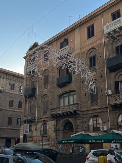 The image depicts a street scene in Palermo, Italy, focusing on the facade of an old building adorned with decorative lights. The building, constructed with stone and featuring multiple stories with arched windows and balconies, is partially obscured by a large, illuminated archway draped across the street. The archway, a festive decoration likely for a holiday or event, is made of a white, intricate metal structure with small, colorful lights.
Below the building, a street is visible with parked cars, including one with a roof box and another in the foreground with a person on a motorcycle next to it. A green awning with "Terzo Cerchio" printed on it stretches across a storefront, indicating a commercial establishment. The sky above is clear and blue. The overall impression is of a lively urban environment, possibly during an evening celebration.