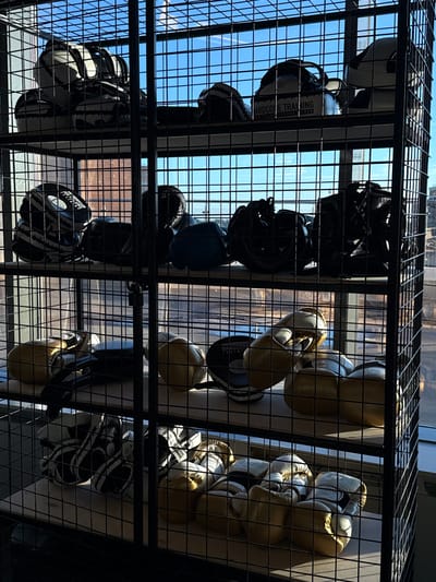 The image depicts a metal shelving unit filled with boxing equipment, including gloves and pads. The unit is positioned in front of a window with a clear blue sky and some clouds visible, suggesting an outdoor view. The lighting indicates it is daytime. The shelves are organized with different types of boxing gear, with some items appearing to be in heavier use than others. The overall setting appears to be an indoor training facility or gym. There are no people visible in the image. The scene is shot from a slightly low angle, looking up at the shelves. The metal grid of the shelving unit creates a patterned effect throughout the image.