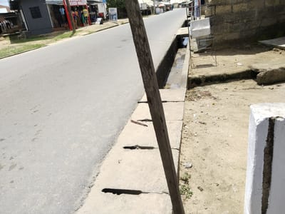 The image depicts a street scene in Uyo, Nigeria. A paved road runs horizontally across the frame. On the left side of the road is a grey building with a sign that reads "SHARON'S FASHION HUB." Mannequins dressed in clothing are visible in front of the store. The right side of the road features a concrete drainage ditch with rectangular openings. A weathered wooden post is positioned near the edge of the ditch, partially obscuring the view. Further down the road, there are more buildings and signs. A metal rack for drying clothes stands between the drainage ditch and a weathered concrete wall. The ground is sandy and there is a white painted concrete block on the right edge of the frame.
