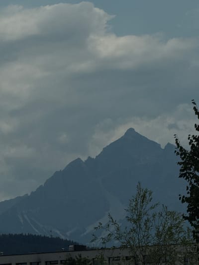 The image depicts a cloudy sky over a mountain range. A building is visible on the right side of the frame, with a tree in the foreground. The lighting suggests it is daytime. The weather appears overcast, with a large cloud mass dominating the left side of the image. The mountain range is dark blue and appears to be covered in vegetation on its lower slopes. The building is modern and has several windows. The tree in the foreground has light green leaves. The overall mood is somewhat somber due to the overcast sky. The location is identified as Innsbruck, Austria.