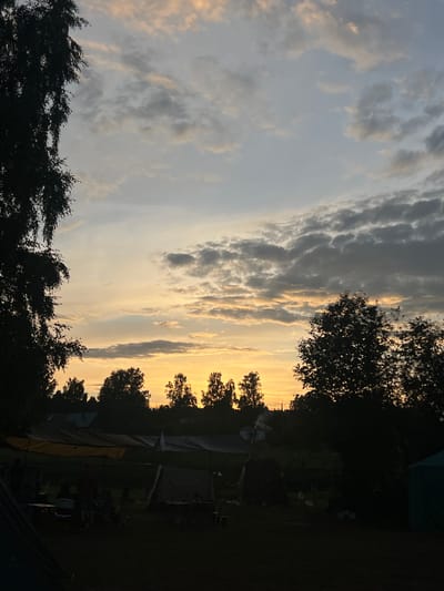 This media file depicts a scene at dusk in Sergino, Russia, likely at a campsite. The sky is filled with dramatic clouds, transitioning from a pale grey on the left to a warm, golden-yellow glow on the right, indicating the setting sun. Silhouetted trees frame the sky on the left and right edges of the image. In the foreground and mid-ground, several tents are set up, suggesting a camping or outdoor gathering. Some figures, appearing as dark silhouettes, are visible near the tents, indicating the presence of people. There is also camping equipment such as chairs and tables scattered around. The overall mood is peaceful and serene, characteristic of an evening outdoors. The time of day is clearly sunset, with the low angle of the sun creating long shadows and a warm color palette in the sky. There is no visible text in the image.