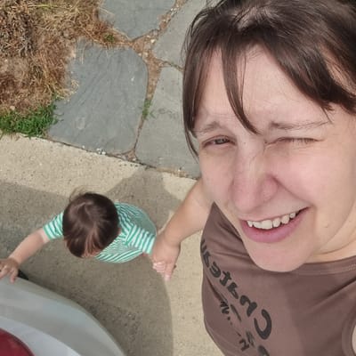 In an outdoor scene, a woman is taking a selfie with a child. The woman is in the foreground, smiling and winking at the camera. She is wearing a brown t-shirt. The child, in the middle ground, is wearing a green and white striped shirt and is holding the woman's hand. The child is looking down. The background shows a paved area with some dry grass and a few small green plants. The scene appears to be in a residential area.