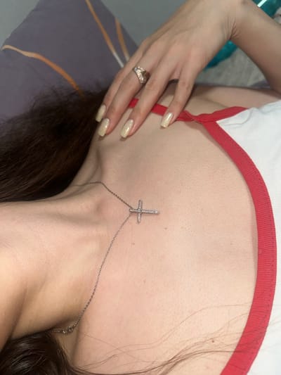 The image shows a close-up view of a person's upper chest, neck, and hand. The person is lying down, possibly on a bed, with their head resting on a pillow. A silver cross necklace is visible on the person's chest, and a ring is worn on the index finger of their hand. The person is wearing a white top with a red trim. The background is blurred and appears to be a room with some colorful objects. There are no discernible landmarks or elements that would indicate a specific location in Postoyata, Ukraine.