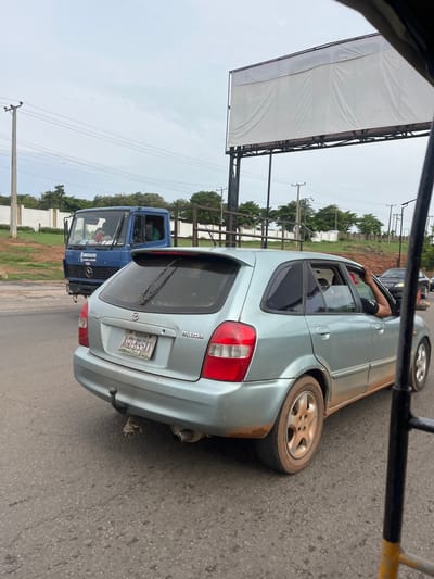 The image displays a daytime roadside scene under an overcast sky, likely in traffic. The primary subject is a light blue/silver Mazda 323F hatchback/wagon, positioned at an angle, with its rear and side visible. A person's arm, wearing a black sleeve or band, is resting outside the open rear passenger window of the Mazda. The lower body of the car appears dirty.
Behind the Mazda, a blue Mercedes-Benz truck is partially visible, featuring the Mercedes-Benz logo on its grille and "Ammisen und Technik" printed on its side. Another dark vehicle is seen further down the road, partially obscured.
To the left, a large, empty white billboard with a dark metal frame stands against the cloudy sky. The setting includes an asphalt road, green trees, fencing, utility poles, and overhead power lines, suggesting a developed area.
Visible text includes "Mazda" and "323F" on the rear of the car. The Mazda's license plate clearly shows "NIGERIA" at the top, "LAGOS" in the middle, and "KJD-393AA" at the bottom. The truck also bears the "Mercedes-Benz" logo and text "Ammisen und Technik". The billboard itself is blank. The overall scene reflects a typical road environment in Nigeria, aligning with the provided context of Kuchingoro.