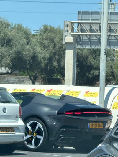 The image captures a scene in what appears to be Herzliya, Israel. A gray car, identified by its license plate "191:00-004", is parked next to a white vehicle with the plate "50:691-38". Roadside advertising is visible with yellow signs bearing Hebrew text and heart shapes. A highway structure and the road marker "53" can be seen in the background, alongside a tree.