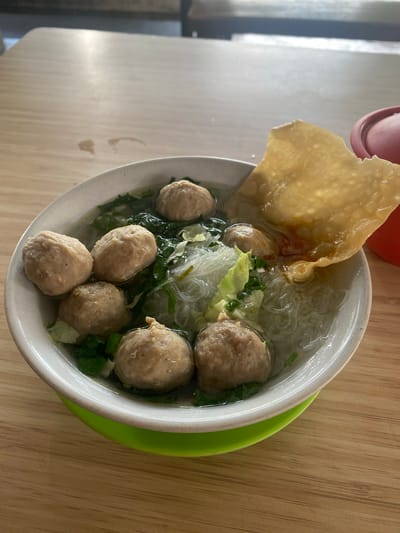A close-up, overhead shot captures a bowl of Indonesian meatballs (bakso) served in broth with noodles and vegetables. The bowl, with its white ceramic interior and light green exterior base, rests on a wooden table. Several round, light brown meatballs are visible submerged in the clear broth, alongside translucent noodles and chopped green leafy vegetables, likely bok choy. A large, golden-brown fried wonton or cracker is perched on the edge of the bowl, partially submerged in the broth, adding a contrasting texture and color. The lighting suggests it is daytime, possibly indoors or in a shaded outdoor area, as indicated by the soft light and lack of harsh shadows. The overall scene evokes a sense of a casual meal, likely in a local eatery in Banjarbaru, Indonesia. No visible text is present in the image.