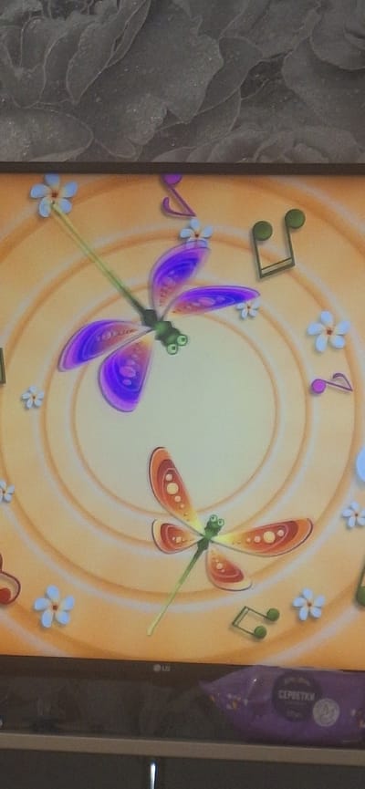 The image depicts a close-up, slightly tilted view of a television screen displaying a cartoonish, whimsical scene. The dominant element is a stylized clock face with concentric rings of a warm, golden-orange hue. Superimposed on this background are two animated dragonflies. The upper dragonfly is predominantly purple with vibrant blue accents on its wings, and it is positioned towards the left side of the screen. The lower dragonfly is orange and yellow with pinkish-red accents, situated towards the bottom right. Both dragonflies have large, green, googly eyes and thin, green bodies. Scattered around the clock face are small, white, five-petaled flowers with light blue centers, and green and pink musical notes. The LG logo is visible on the bottom edge of the television screen, indicating the manufacturer. Below the screen, a purple package with text that appears to be in Cyrillic script is partially visible. The scene is illuminated by ambient lighting, casting a soft glow. The background of the actual room, above the television, is a textured gray surface resembling stone or concrete. There are no people or identifiable landmarks of Bila Tserkva, Ukraine visible in the image.