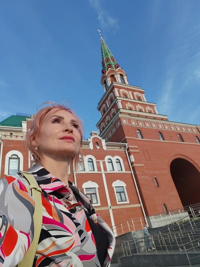 A woman with pink hair stands in front of a large, red brick building with a tall, green-capped tower. She is looking up and to the right, with a slight smile and her eyes closed. She is wearing a colorful, patterned shirt and a light-colored strap over her shoulder. The building appears to be a historic or ornate structure, with multiple windows and decorative white trim. The sky is clear and blue with a few wispy clouds. The sun is bright, suggesting it is daytime. The woman's expression and the bright lighting suggest a pleasant and possibly reflective mood. In the background, there are stairs with railings leading up to an arched entrance of the building. A person in a light blue outfit can be seen walking up the stairs. The overall scene appears to be a tourist or recreational setting, possibly in a city square or historical site.