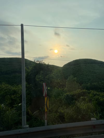 The image depicts a landscape in Aê Thi, Vietnam, under a hazy, bright sky with the sun visible. A lush, green hillside covered in vegetation occupies the right half of the frame. In the foreground, a weathered grey metal barrier with red, yellow, and white reflective stripes is present. Also in the foreground, an unlabeled, gray metal pole bisects the view. Power lines run horizontally across the sky and hillside. The lighting suggests either sunrise or sunset. No people are visible.