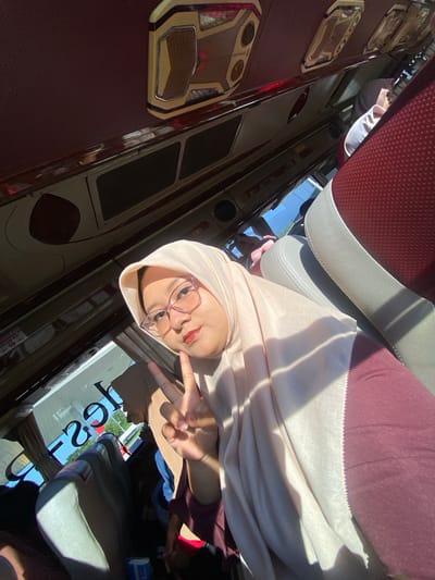 The image depicts a female individual taking a selfie inside a passenger bus or coach, under bright daylight conditions. The main subject, positioned in the foreground, is an East Asian or Southeast Asian woman wearing a light peach-colored hijab and what appears to be dark red or maroon clothing. She has purple-framed eyeglasses and a reddish-orange tint on her lips. Her right hand is raised, forming a "V" or peace sign, positioned near her chin, as she looks directly at the camera.

The interior of the bus features seats upholstered in a combination of dark red/maroon textured fabric and light grey/white smooth material. The ceiling is predominantly dark red, fitted with light-colored, rectangular overhead service units that include lighting elements and metallic trim. A reflective strip or handrail runs along the upper window line. Sunlight streams into the bus from the side, creating distinct shadows on the main subject's face and hijab.

Behind the main subject, another individual wearing a brown hijab is partially visible, seated further back in the bus. Through the windows, a bright blue sky and green foliage are visible, indicating an outdoor setting, possibly alongside a road or at a stop. A reversed reflection of "26s" or similar characters is discernible on the front window, suggesting an external sign or vehicle. The overall scene is consistent with a journey on a passenger coach. While no specific visual markers identify the location within the image, the scene is situated within a region consistent with Sumber Suko, Indonesia.