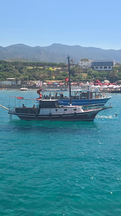 The image depicts a bright, sunny day at a coastal location, likely Karavas, Northern Cyprus. Two boats are prominent in the turquoise sea. The foremost boat is a traditional wooden vessel with a dark red hull and a white upper deck, carrying several passengers. The name "SERNA" is visible on its bow. Behind it, a larger, blue-hulled boat is also present, with more passengers visible on its deck. The background reveals a picturesque coastline with lush green hills and mountains. Buildings with white facades and modern architecture are nestled amongst trees. A beach area with numerous red and white umbrellas suggests a popular tourist spot. The water is clear and calm, with sunlight glinting off its surface. A Turkish flag is visible on a mast of one of the boats.