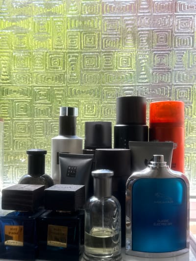 A still life image showcases a collection of men's grooming products and perfumes arranged on a white surface in front of a textured, green-tinted glass window. The window features a repeating pattern of concentric squares. The foreground displays two rectangular blue glass perfume bottles, each with a dark wooden cap and a gold label. The left bottle is labeled "NUIT D'AZAR" and "RITUALS...", while the right is labeled "BLEU BYZANTIN" and "RITUALS...". To the right of these is a clear glass perfume bottle with a silver spray top, containing a light yellow liquid, and further to the right is a large, blue, glass perfume bottle with a silver rim and cap, featuring the Jaguar logo and the text "CLASSIC ELECTRIC SKY". Behind these, a variety of other bottles and tubes are visible, including a white bottle with a black cap, a gray tube, several dark, cylindrical containers, and a bright orange aerosol can. The lighting suggests it is daytime, with natural light filtering through the patterned glass. The scene appears to be indoors, possibly on a windowsill or shelf in a bathroom or dressing area. The arrangement of items suggests they are personal care products. The location is identified as Oeiras, Portugal.