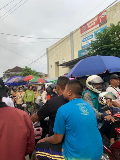 The scene is a crowded street in Kecamatan Lawang, Indonesia, located at coordinates -7.848 latitude, 112.699 longitude. The street is lined with shops, including one with a large sign that says "NIPPON PAINT". Several people are crowded around the street. A man in a blue t-shirt is standing in the foreground, while several other people are visible in the background. Some of the people are holding umbrellas. A motorbike is parked in the foreground. The sky is cloudy. The people appear to be waiting in line for something. They are interacting with each other by talking and looking at each other. The overall scene is crowded and busy.