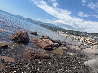The image depicts a rocky beach in Semidvorye, Ukraine, where the shoreline meets the sea. Large, reddish-brown rocks are scattered along the edge, partially submerged in the water. Smaller, dark pebbles cover the beach, with some appearing wet and shiny. In the distance, buildings and trees line the coast, and mountains rise in the background. The sky is blue with scattered white clouds. The water is clear and reflects the sky.