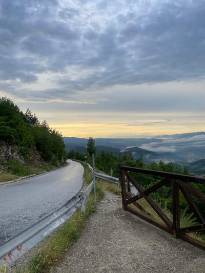 The image depicts a scenic mountain landscape at dawn or dusk. A paved, winding road curves through dense green foliage and rocky terrain, bordered by a metal guardrail. In the foreground, a rustic wooden gate stands open, suggesting access to a viewpoint or a trail. The sky is filled with dramatic clouds, ranging from dark gray to lighter tones, with a band of soft yellow light on the horizon, indicating either sunrise or sunset. In the distance, layered mountain ranges are visible, partially obscured by wisps of mist, creating a sense of depth and tranquility. The overall mood is peaceful and awe-inspiring, highlighting the natural beauty of the mountainous region. No visible text is present in the image. The location context is Delchevo, Bulgaria.