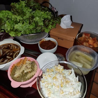 A close-up, eye-level shot captures a spread of Korean side dishes and pasta arranged on a dark wooden table. The primary focus is on a large metal colander overflowing with vibrant green and burgundy lettuce leaves, positioned on the left side of the frame. To the right of the lettuce is a small, light brown rectangular box with Korean text printed on it. Adjacent to the box, a white paper napkin is folded and resting on its edge.
Below the lettuce, a white oval dish contains a dark, savory broth with sliced onions. Next to this dish is a small white bowl filled with a reddish-brown paste, likely a spicy condiment. In the foreground, a pink plastic bowl holds tri-colored fusilli pasta. To the right of the pasta, a clear glass bowl contains a creamy coleslaw mixed with corn kernels. Further to the right, another clear plastic container holds pickled radishes. A small dish of kimchi is visible in the upper right corner. Chopsticks and a spoon are also present on the table. The lighting is dim, suggesting an indoor setting, likely a mealtime. The scene is set in Dunnae-myeon, South Korea.