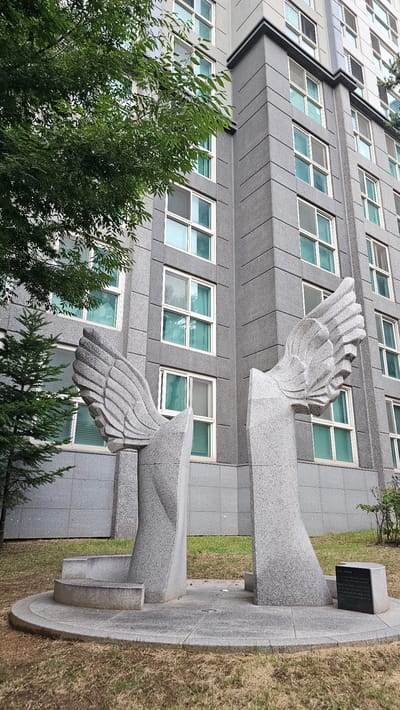 This image depicts a public art installation featuring two large, abstract sculptures resembling wings, made of granite. The sculptures are positioned in front of a modern, multi-story building with a grey, textured facade and numerous windows. Green trees and grass surround the sculptures and building, suggesting an outdoor urban setting. A small, black plaque with Korean text is visible on the ground to the right of the sculptures. The setting appears to be a communal space or park area within a residential complex. The art installation is located in Asan-si, South Korea.