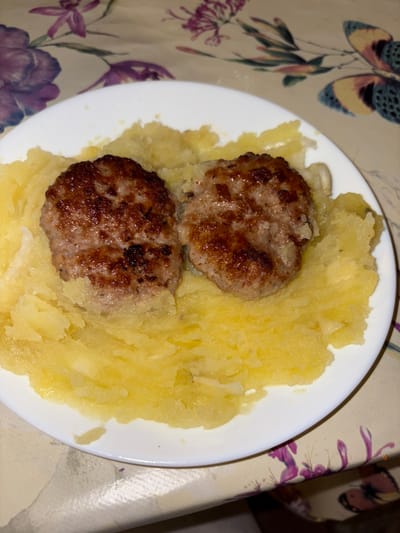 This image captures a meal served on a white, circular ceramic plate, situated on a patterned tablecloth. The scene is located in Batoshevo, Bulgaria. The plate contains two oval-shaped, pan-fried meat patties, characterized by browned, textured surfaces, positioned side-by-side near the center. These patties rest upon a substantial bed of yellow, coarsely mashed or pureed potatoes, which fills the remainder of the plate. The potatoes exhibit a lumpy consistency, with small, lighter-colored chunks visible throughout. The tablecloth features a multi-colored design incorporating purple and pink floral motifs, alongside yellow, blue, and brown butterfly patterns. No individuals or active interactions are depicted. The lighting suggests an indoor setting.