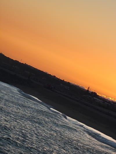 A diagonal shot captures the ocean meeting the shore at Quarteira, Portugal, during sunset. The sky is a gradient of warm orange and yellow, with faint clouds near the horizon. The ocean is dark blue with white foamy waves lapping against the dark sandy beach. Silhouetted against the sunset are a few people walking along the shoreline and some structures indicating a coastal town. The scene has a serene, end-of-day atmosphere.