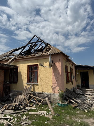 The image displays a single-story residential house in Nedryhailiv, Ukraine, exhibiting significant damage under a partly cloudy sky. The roof structure is severely compromised, with numerous charred and broken wooden rafters exposed and much of the grey corrugated roofing material dislodged or missing. The house exterior features textured yellow and peach-colored stucco walls above a stone or concrete foundation. Two prominent front windows with brown frames have shattered glass panes. A white satellite dish is mounted on the yellow wall. Scattered across the green grass and against the house are extensive debris piles, consisting of broken grey roofing fragments, charred wooden beams, and a dismantled wooden window frame. A green plastic planter with a small yellow flowering plant is positioned near the debris on the right. An open doorway on the right side of the house reveals the lower legs of a person wearing shorts within the interior. Electrical wiring runs along the exterior walls.