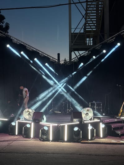 An outdoor stage at dusk in Busmantsi, Bulgaria, is illuminated by dramatic crisscrossing spotlights. A man wearing a white t-shirt and tan pants stands center stage, holding a microphone stand. In the background, a drum set and various audio equipment are visible. Several large industrial fans are placed around the stage. The sky is a deep blue, transitioning to a lighter hue near the horizon. A metal structure with stairs is situated behind the stage, suggesting an industrial or event venue setting. The foreground shows a rough, paved surface.