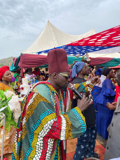 The image depicts an outdoor event, possibly a wedding or celebration, in Kuru Dungus, Nigeria. A man in a colorful patterned shirt and maroon fez is speaking into a microphone. The background reveals a crowd of people under a canopy adorned with red, white, and blue fabric, as well as maroon and green drapes. There's a tiered white cake visible in the background. The sky appears overcast. People are gathered, seemingly listening to the speaker. In the foreground are white flowers.