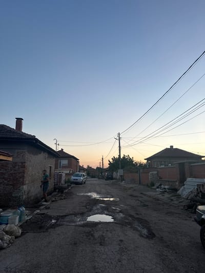 The image depicts a residential street scene in Malevo, Bulgaria, at dusk. The sky is a gradient of pale blue to soft orange, suggesting the sun is setting. Power lines crisscross the sky above a row of brick houses with tiled roofs. The street is unpaved and muddy, with several large puddles reflecting the dim light. On the right side of the street, a man is performing a push-up next to a collection of plastic water bottles and bags. A silver car is parked further down the street, and another vehicle, possibly a van, is partially visible near the houses on the left. The overall impression is one of a rural or semi-rural setting in the early evening, with a casual, everyday activity taking place amidst the somewhat disheveled environment.