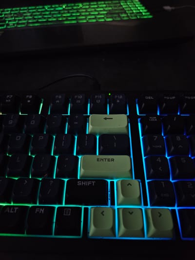 The image displays a close-up view of a black mechanical keyboard with RGB backlighting. The lighting is a mix of green and blue, with the top row of keys and the keys on the left side illuminated in green, and the right side and bottom rows in blue. Several keys are clearly visible and identifiable. In the top row, function keys such as F7, F8, F9, F10, F11, and F12 are present. Below these, the number keys 9 and 0 are visible, along with the equals sign key. Further down, the letter keys O and P are present, as well as bracket keys. On the left side of the keyboard, the Shift key and the Alt and Fn keys are visible. The Enter key, a light green color, is prominently featured. The arrow keys, also light green, are visible in the lower right quadrant of the image. The top of a second keyboard, also with green backlighting, is visible at the very top of the frame. There are no people or indications of a specific location within the city of Versailles, France.
