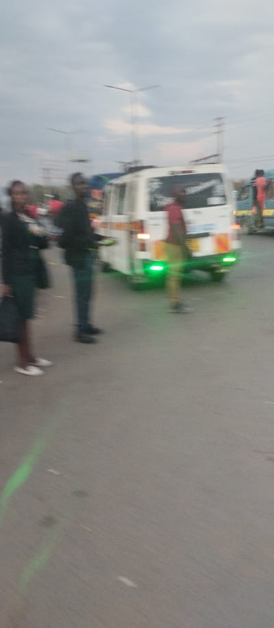 The image shows a street scene in Kisumu, Kenya (latitude: -0.091, longitude: 34.769). There are three individuals present: two adults and one child. Two adults are in the foreground, both are facing towards the camera, and one adult is out of focus. The adults appear to be in their twenties. There is a child, also out of focus, walking behind a white minivan. The white minivan has a yellow stripe along the side. The scene is set on an asphalt road. The weather is overcast and the sky is grey. There is a street lamp in the background. The white minivan has a green light on the back.